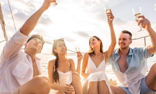 Group of diverse friends drink champagne while having a party in yacht. Attractive young men and women hanging out, celebrating holiday vacation trip while catamaran boat sailing during summer sunset.